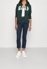Felpa verde con zip e lettering bianco "G.A.P.", abbinata a jeans cropped in denim scuro e sneakers bianche. Tessuto morbido e design aderente.