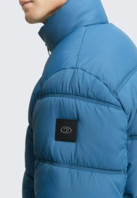 Plavi puffer jakna s visokim ovratnikom, s crnom logotip oznakom na rukavu, kviltanom teksturom i glatkom, sjajnom završnom obradom.