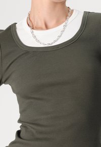 Top a maniche lunghe verde oliva a costine sovrapposto a una maglietta bianca a costine con scollo rotondo, presenta un collo a fondo scollato e una collana in argento.