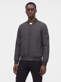 Uomo di colore che indossa una giacca bomber grigio scuro con cerniera frontale e tasche laterali, in piedi davanti a uno sfondo grigio chiaro uniforme.