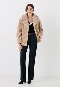 Abrigo de piel sintética beige con un amplio cuello, combinado con una blusa negra de escote en V con rayas blancas y rojas, y pantalones vaqueros oscuros. Botas de tacón negras.