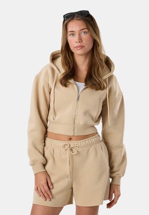 Young woman with long hair wearing beige hooded zip-up jacket and matching shorts, sunglasses on head, standing neutral against white background.