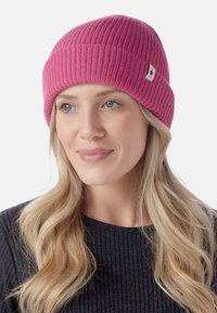 Mujer con cabello rubio largo que lleva un gorro de punto rosa y un suéter negro de canalé, mirando ligeramente de lado con una expresión neutral.