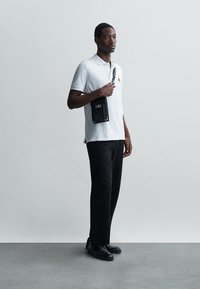 Vit pikétröja med en liten broderad design, svarta byxor och svarta skor. Personen håller en svart axelremsväska.