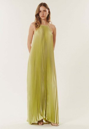 Femme portant une robe maxi sans manches, plissée, vert citron à col licou, avec des talons ouverts dorés, devant un fond uni.