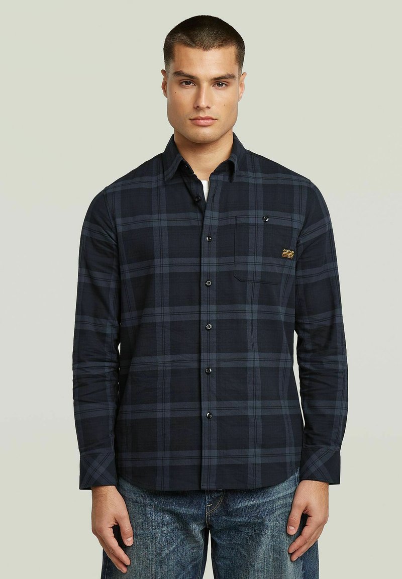 Camicia a maniche lunghe a quadri blu navy e grigia con chiusura a bottoni, tasca sul petto, colletto e tessuto di cotone testurizzato, indossata con jeans blu.