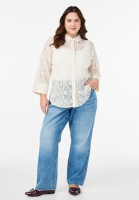 Koszula z koronką zapinana na guziki w odcieniu kremowym, z kołnierzykiem, długimi rękawami i kieszonką z przodu. W połączeniu z jasnoniebieskimi dżinsami o luźnym kroju i ciemnymi butami.