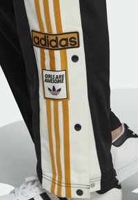 Svarta och vita Adidas-byxor med gula ränder, tryckknappar, märken med texten "adidas" och "GIRLS ARE AWESOME," buren med vita Adidas-sneakers.