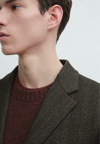 Blazer marrone a spina di pesce con rever a scialle, abbinato a un maglione bordeaux con motivo a rilievo. Primo piano del colletto e della spalla.