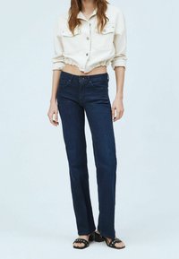 Korkeavyötäröiset tummansiniset farkut, joissa on suora lahje. Yhdistettynä lyhytvartiseen beigeen napitettuun takkiin ja mustiin korkokenkiin.