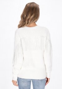 Maglione bianco con dettagli in frange argento sul retro, texture a maglia, collo rotondo e orlo a coste. Abbinato a jeans azzurri chiari.