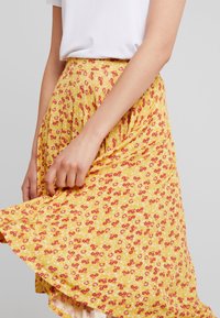 Keltainen kukkakuvioinen hame, jossa punaisia ja valkoisia kuvioita, kevyt kangas, laskostettu malli, polven pituinen ja laskeutuva helma.