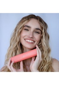 Spray Curl Curl dans un flacon cylindrique rose corail avec du texte blanc, tenu par une personne aux cheveux ondulés et au sourire éclatant sur un fond clair.