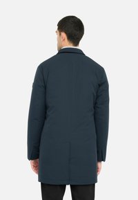 Uomo che indossa un lungo cappotto blu scuro, in piedi con la schiena rivolta verso la telecamera su uno sfondo bianco semplice.