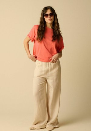 Frau mit langen welligen Haaren, die runde Sonnenbrille, ein korallenfarbenes kurzärmeliges Hemd, weite beige Hosen und gewebte Slip-On-Schuhe vor einem schlichten Hintergrund trägt.