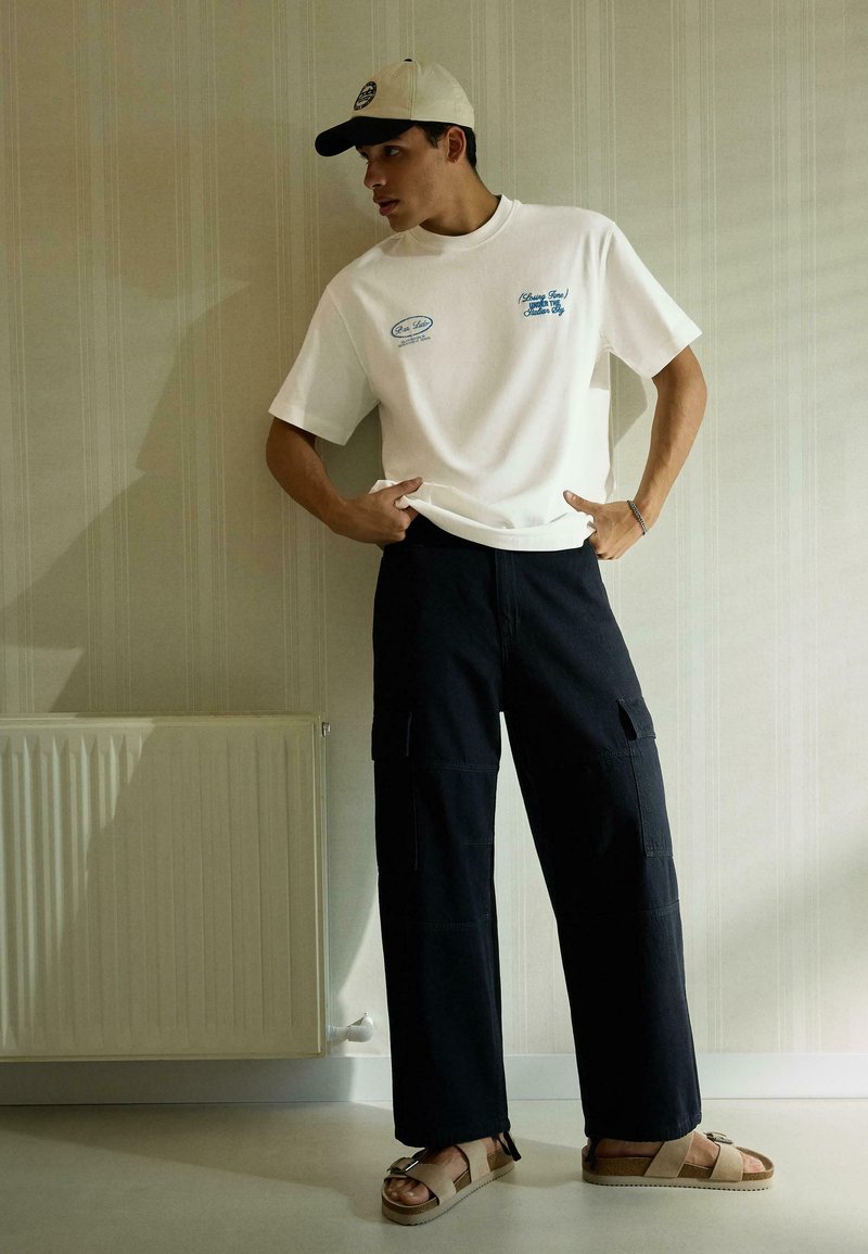 Young man wearing beige cap, white graphic t-shirt, black cargo pants, and beige sandals standing in a room with a radiator and striped wallpaper.
