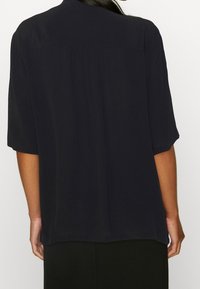 Schwarze Bluse mit kurzen Ärmeln und lockerer Passform, die mit einem gefältelten Rückendetail und glatter Textur versehen ist. Keine sichtbaren Muster oder Beschläge.