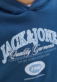 Hoodie bleu en coton avec une texture douce. Présente un texte imprimé en blanc : "JACK&JONES Quality Garments" et un logo rond avec "1990."