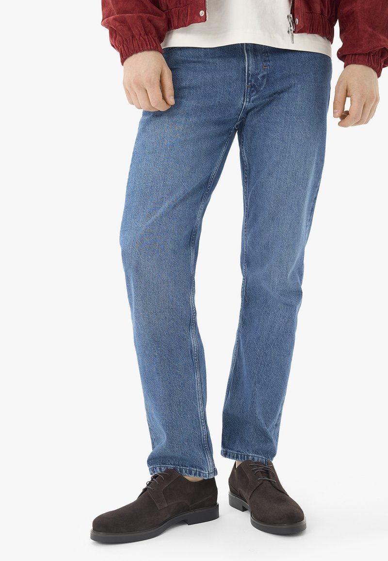 Homme portant un jean bleu, des chaussures marron foncé à lacets, une chemise blanche et une veste rouge, debout avec les mains le long du corps.