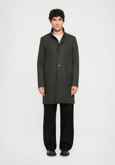 Manteau en laine gris foncé avec un col haut et une fermeture à un seul bouton, comportant deux poches latérales et une coupe ajustée. Associé à un pantalon noir à jambes larges.