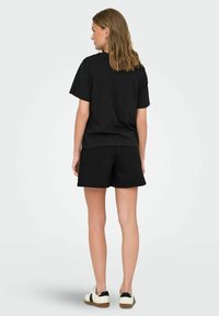 Schwarzes Baumwoll-T-Shirt mit kurzen Ärmeln, kombiniert mit schwarzen Shorts. Das Outfit wird durch weiße Sneaker mit schwarzen Akzenten abgerundet.