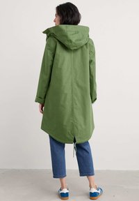 Veste à capuche verte avec une coupe longue et droite, dotée d'un cordon de serrage à l'ourlet arrière. Portée par-dessus un jean bleu court et des baskets bleues.