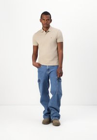 Beige polo tričko s krátkým rukávem a texturovaným pletením, spárované s modrými cargo kalhotami s kapsami. Hnědé boty dokončují outfit.