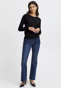 Cardigan negru tricotat cu decolteu rotund, închidere cu nasturi și mâneci lungi, asortat cu blugi albaștri cu croială dreaptă și mules negre cu toc.