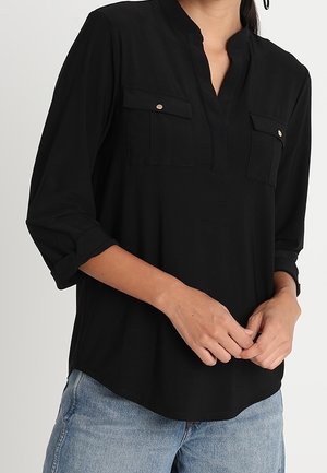 Vrouw draagt een zwarte blouse met lange mouwen en borstzakken en een blauwe spijkerbroek, handen losjes in elkaar gevouwen ter hoogte van de taille.