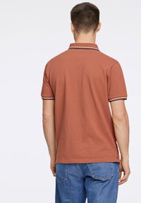 Polo shirt de couleur rouille avec un tissu texturé, manches courtes et accents de rayures foncées sur le col. Porté avec un pantalon en denim bleu.