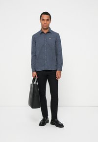 Camicia a fantasia checked navy con collarino button-down, pantaloni slim-fit neri e borsa nera a spalla. Tessuto morbido, taglio casual, hardware minimale.