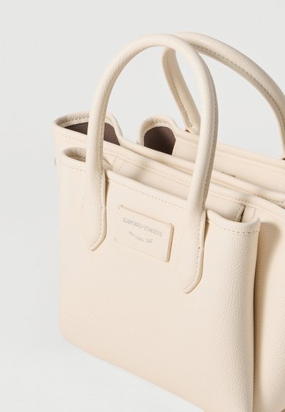 Bolso de mano de cuero crema con asas dobles en la parte superior, superficie texturizada y una sutil placa con logo. Forma rectangular compacta con apertura en la parte superior.
