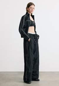 Γυναίκα που στέκεται φορώντας μαύρο αθλητικό σύνολο με λευκές ρίγες, τα χέρια στις τσέπες, με κοντό τοπ, φαρδιά φόρμα και αθλητικά παπούτσια σε γκρι πάτωμα.