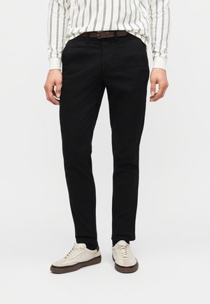 Muž v úzkych čiernych nohaviciach, hnedom opasku, bielej pruhovanej košeli a béžových teniskách stojaci pred jednoduchým svetlým pozadím.