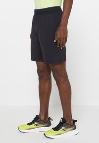 Czarne sportowe szorty z elastycznym pasem, bocznymi kieszeniami i luźnym krojem; zestawione z zielono-czarnymi butami do biegania.