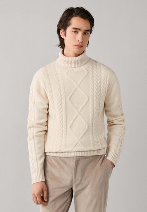Junger Mann mit dunklen Haaren, der einen cremefarbenen, grob gestrickten Rollkragenpullover und beige Cordhosen trägt, steht vor einem einfarbigen Hintergrund.