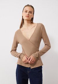 Beige geripptes Langarmtop mit tiefem V-Ausschnitt und Überlappungsdesign, ausgestattet mit detaillierten Manschetten und einer taillierten Silhouette. Kombiniert mit dunkler Jeans.