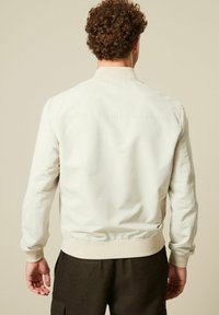 Lys beige bomberjakke med ribbet manchet og talje. Glat stof, minimale sømdetaljer og et klassisk krave-design.