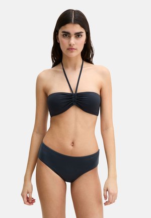 Młoda kobieta o długich ciemnych włosach, ubrana w czarne dwuczęściowe bikini z wiązaniem na szyi, stojąca na jasnym, jednolitym tle.