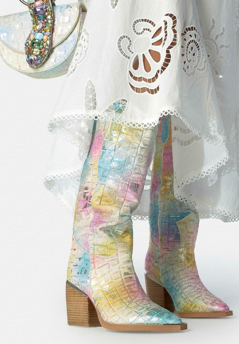 Stivali al ginocchio con texture in coccodrillo pastello multicolore e tacchi in legno impilati abbinati a un vestito bianco con motivi traforati in pizzo.