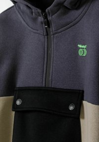 Sweatshirt à capuche avec un zip sur le devant, en tissu gris foncé, avec des blocs de couleurs beige et noir, un petit logo vert et des détails à pression.