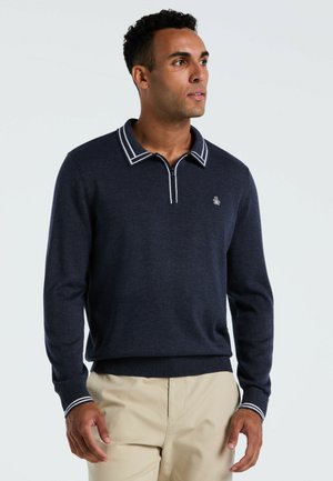 Man in een marineblauwe lange mouwen pullover met een witgestreepte kraag en manchetten, gecombineerd met beige broek, kijkend opzij.