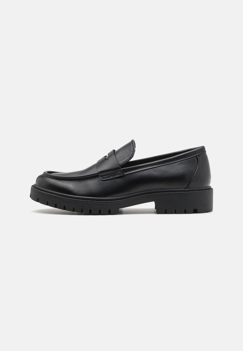 Mocassin en cuir noir avec semelle crantée, petit talon, et une bride à l'avant, vu de profil sur un fond uni.