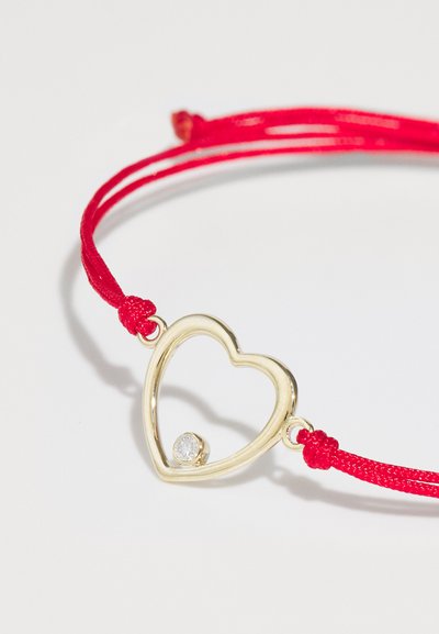 Zelta sirds formas aproce ar centrālu caurspīdīgu akmeni, piekārta uz sarkana aukliņa. Iekļauj slidenu mezglu, lai pielāgotu izmēru.