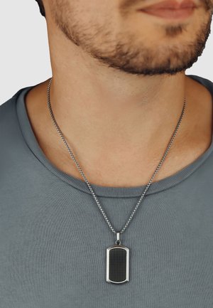 Silberne Kettenhalskette mit einem rechteckigen Anhänger, der eine schwarze strukturierte Oberfläche und einen glänzenden Silberrand aufweist. Getragen auf einem grauen Hemd.