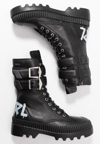 Botas de combate de cuero negro con hebillas plateadas, una cremallera lateral y suelas de goma texturizadas que presentan un prominente logo azul en el lateral.