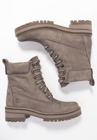 Grå läderankelboots har en texturerad yta, snörning fram och en kraftig gummisula med markanta mönster. Timberland-logotyp synlig.