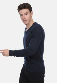 Jeune homme portant un pull V en bleu marine et un pantalon noir, marchant, regardant légèrement sur le côté, sur un fond blanc uni.