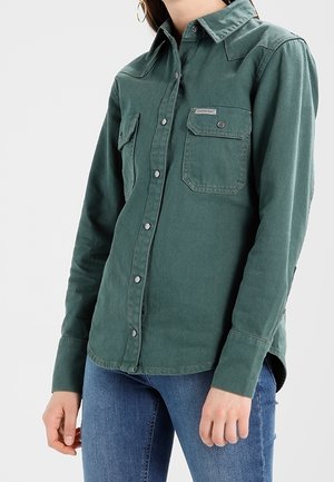 Žena v zelené košili na knoflíky s kapsami na prsou a modrých džínách, zobrazená od krku po boky na jednobarevném pozadí.