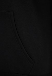 Musta puuvillapaita, jossa on etutasku, sileä pinta ja vahvistetut saumat helmassa. Läheltä katsottuna taskun yksityiskohta.